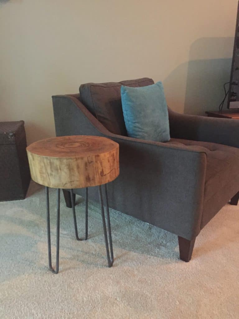 A tree slice side table next to a dark gray chair sitting on beige carpet.