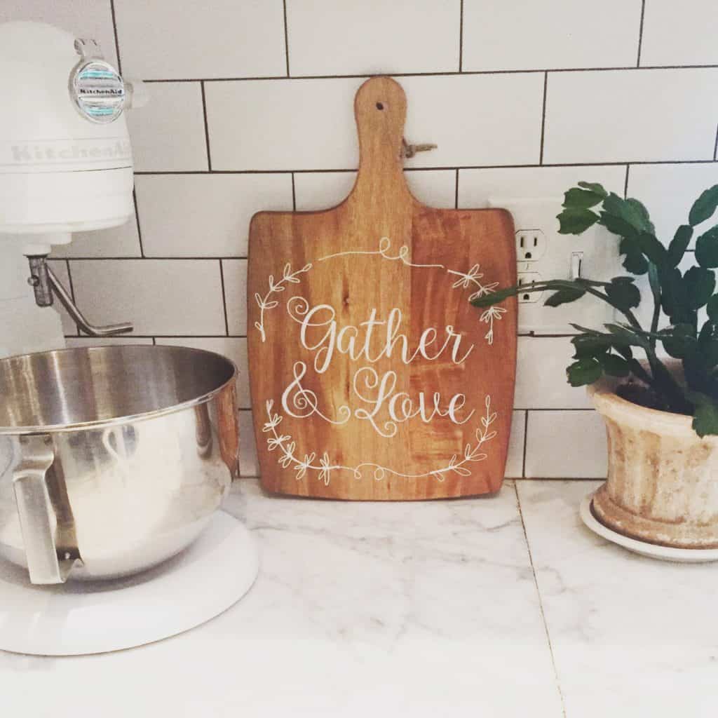 A cutting board that says gather and love that I bought at Tuesday Morning leaning agains my white subway tile backsplash.