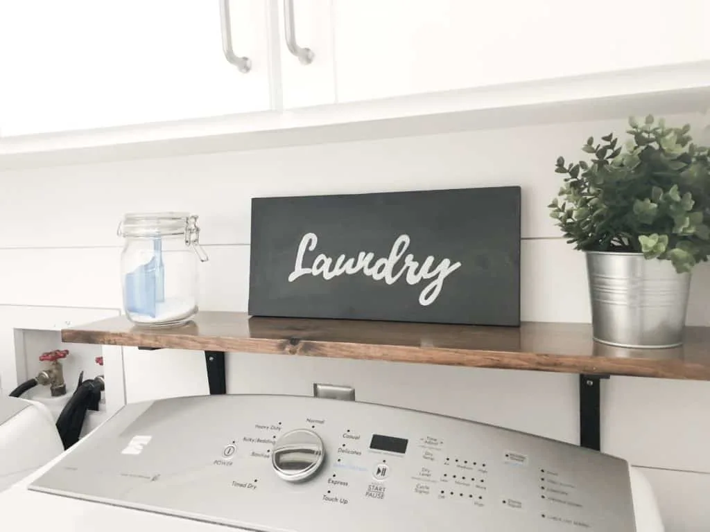 This laundry sign sits on a wood shelf over the dryer with a galvanized bucket and faux plant in it.