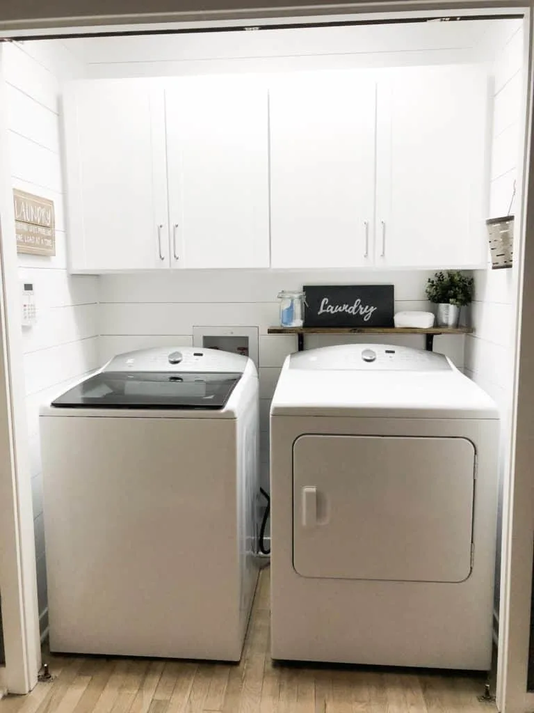 This shows my while farmhouse style laundry room with the white cabinets, white shiplap, and shelf over the dryer.