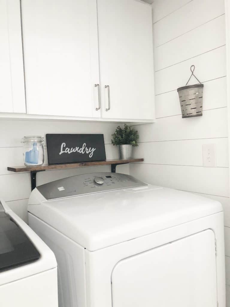 A laundry room with white shiplap walls, a shelf over the dryer with a sign, plant, and laundry detergent.
