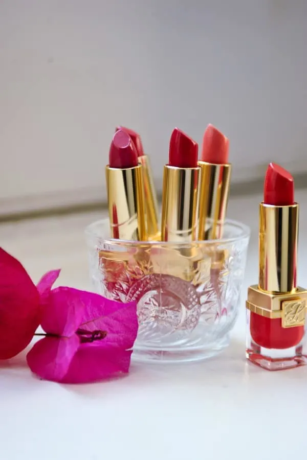 A glass container holding lipsticks and a dark pink flower laying next to it.