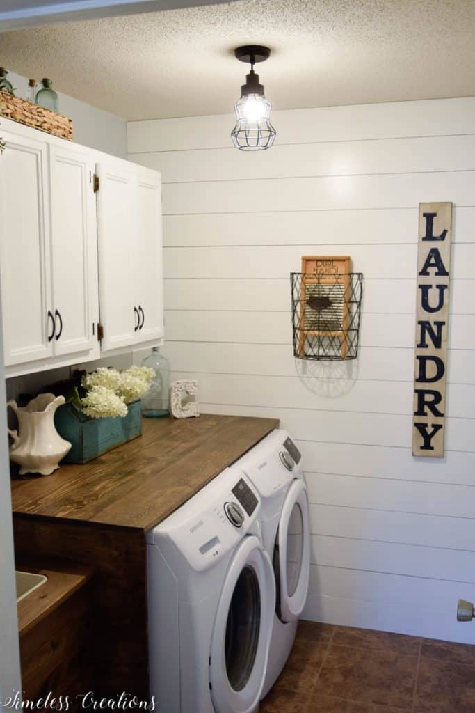 This laundry room has white cabinets above a wood topped washer and dryer and shiplap on the perpendicular wall.