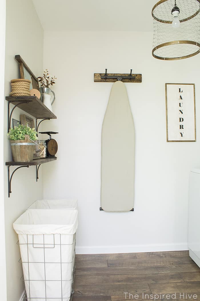 This laundry room has white painted walls with wood floors and an ironing board hanging on the wall next to a laundry farmhouse sign.