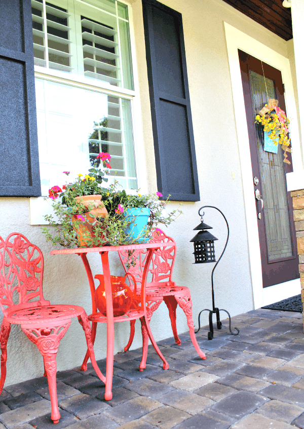 Wrought Iron Chairs that are painted bright pink with flowers on top next to a lantern on a paver patio.