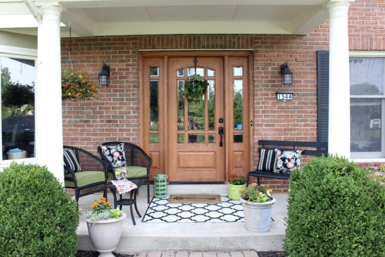 A front porch with black chairs and green cushions and a geometric rug with lots of pots with flowers.