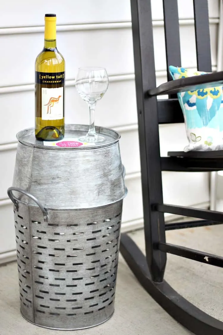 A black rocking chair with a galvanized olive basket next to it with a bottle of wine and a glass.