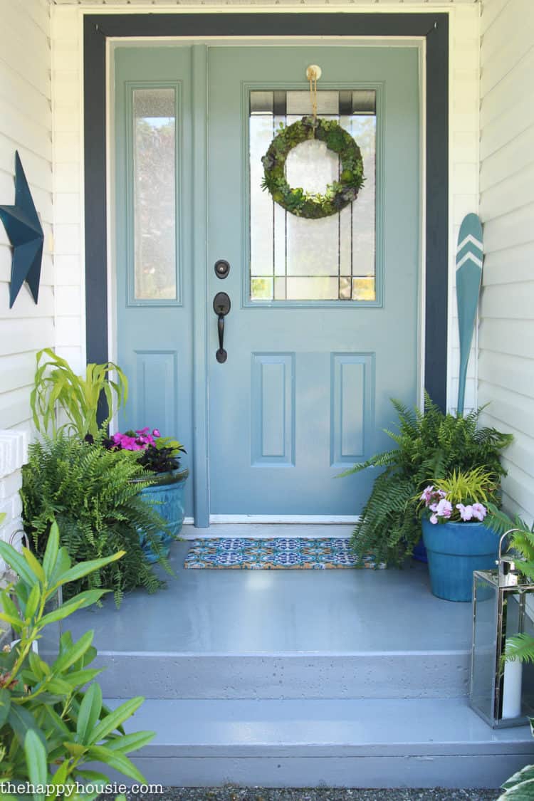 A small front porch with a light blue door and colorful rug with ferns and purple flowers.