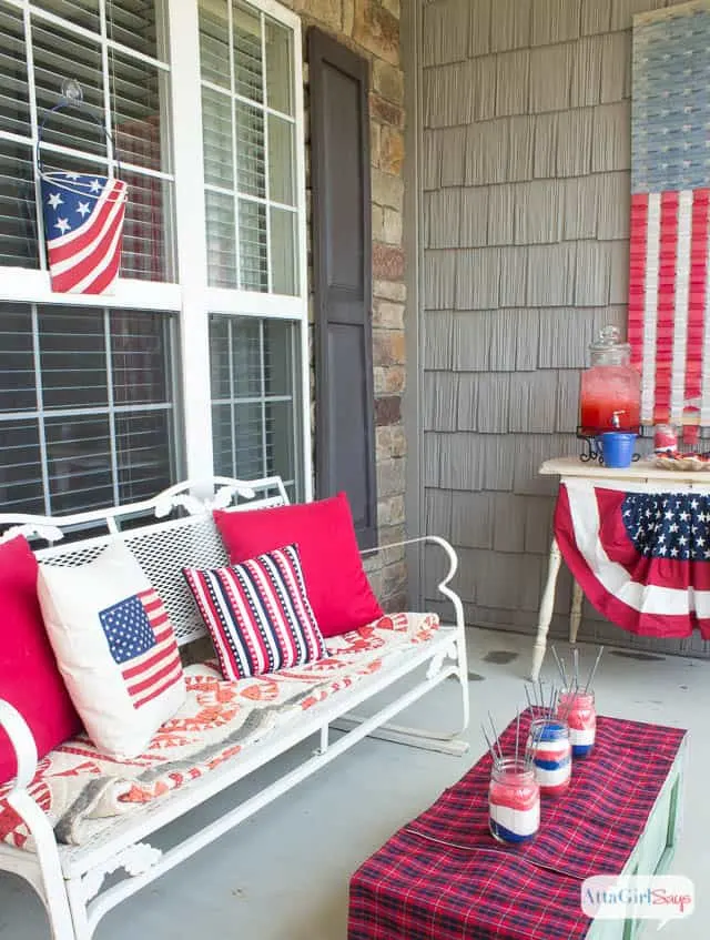 A patriotic front porch with lots of red white and blue in the pillows on the white bench and bunting on the console table.