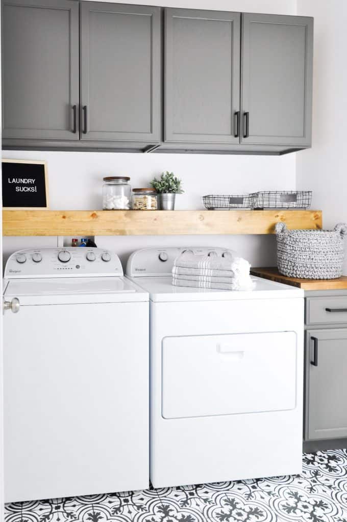 This laundry room has gray painted cabinets and a wood shelf above the washer dryer and a farmhouse sign that says laundry sucks.