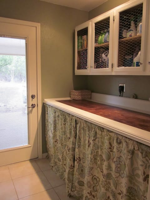 This laundry room has a wood counter with a fabric curtain hiding the machines.  The cabinet doors have chicken wire for fronts.