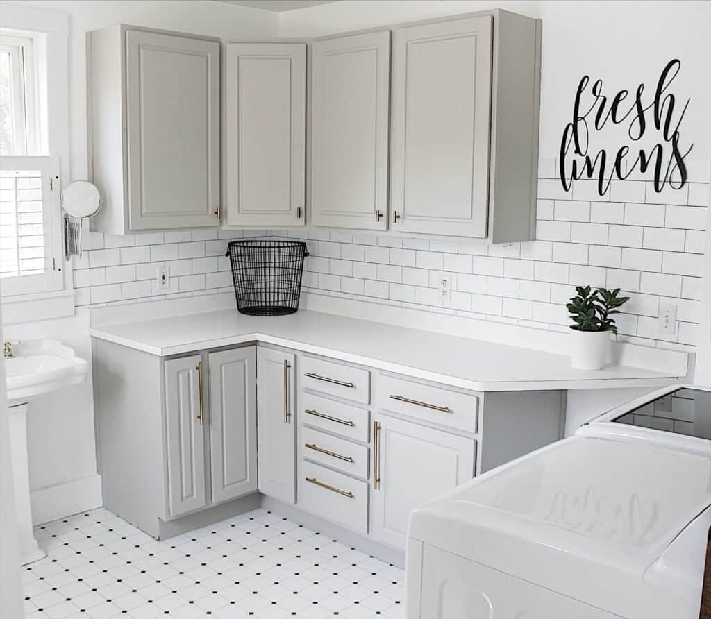 This laundry room has white walls, white subway tile, white counters and gray cabinets.