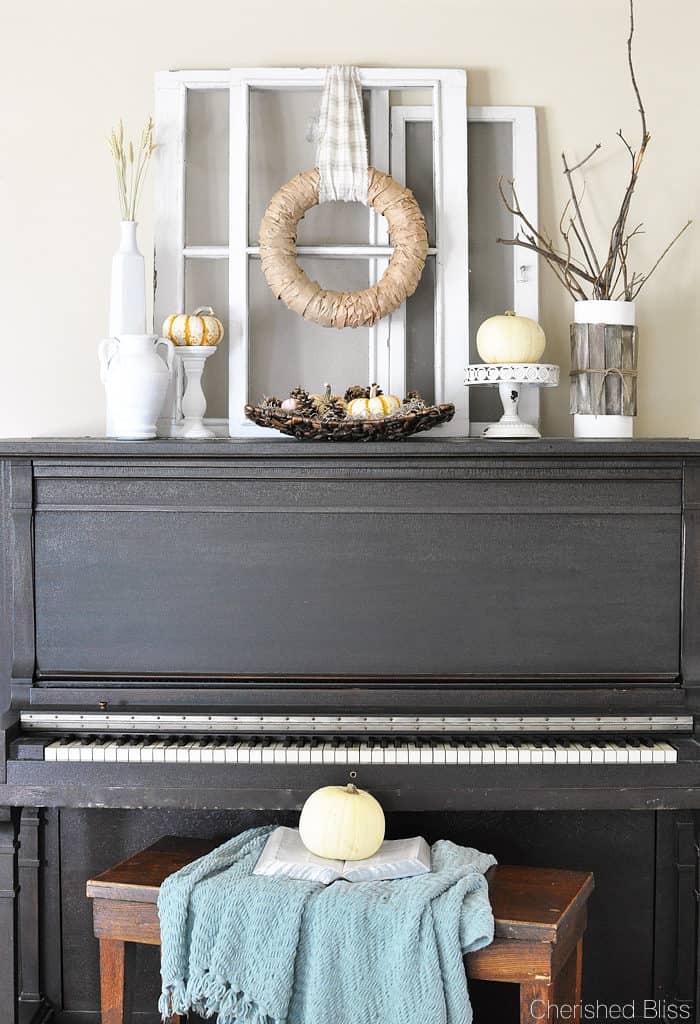 A black upright piano with the top decorated for fall with a couple old windows, a neutral wreath and lots of white with pumpkins.