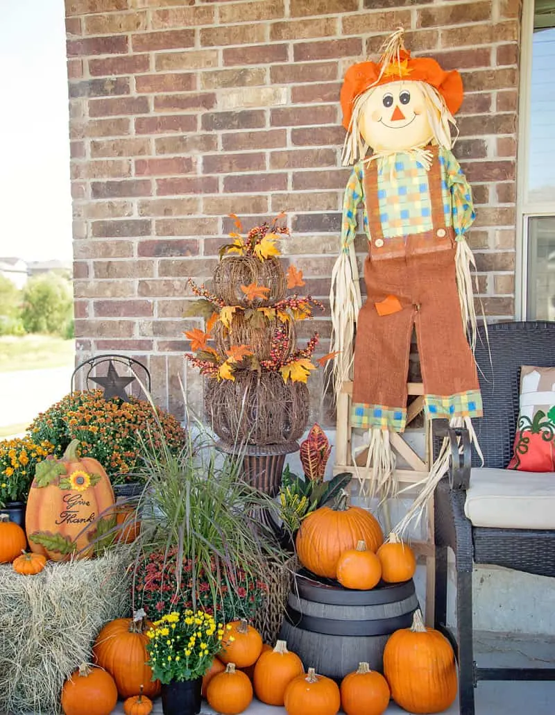 A front porch with a scarecrow, orange leaves and pumpkins and mums in baskets.