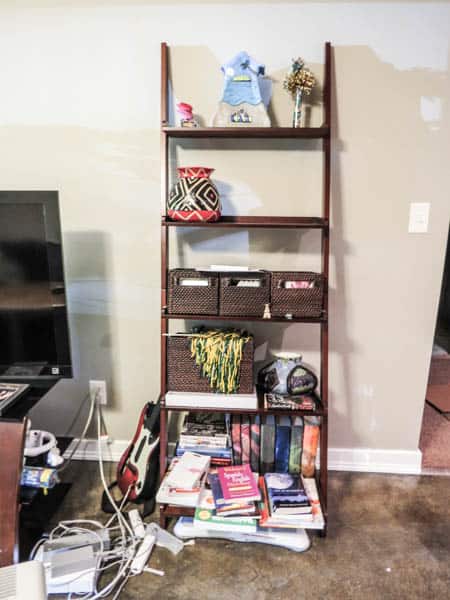 A leaning shelf unit that is a hodge podge of stuff and spilling out to the floorl.