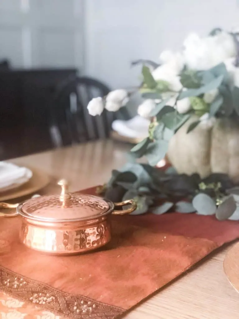 A copper dish on a copper and gold runner next to a pumpkin flower arrangement.