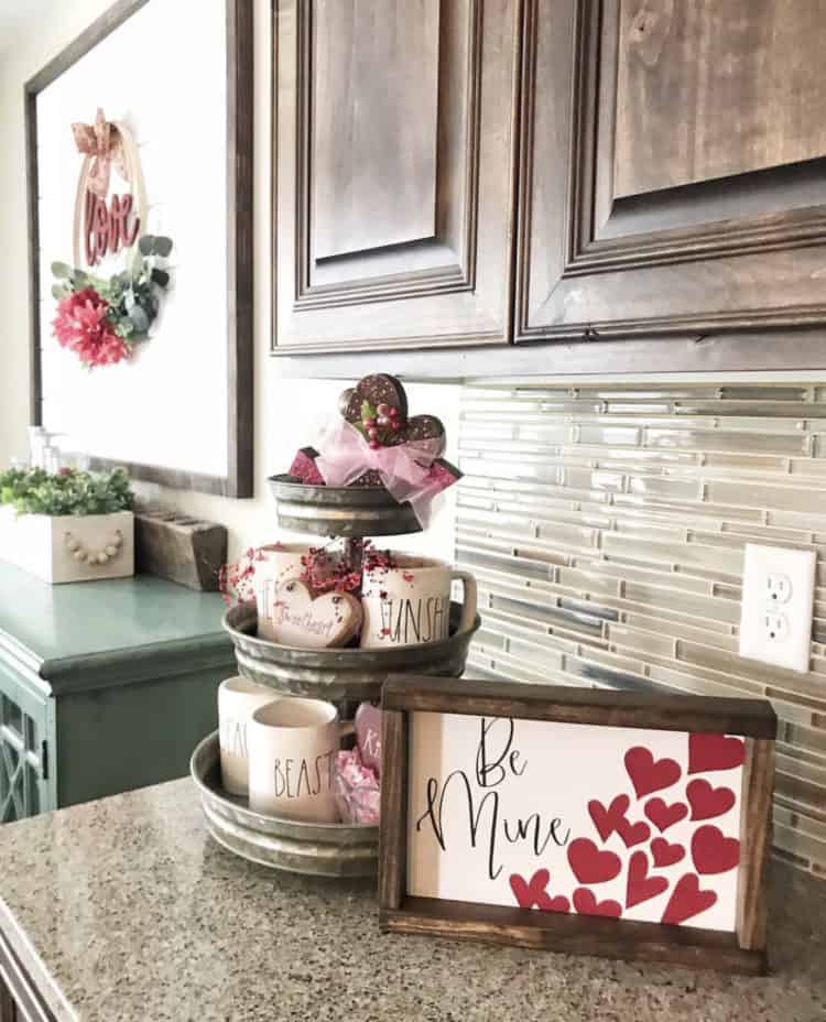 A pretty farmhouse sign that says be mine with hearts on it next to a tiered tray with Rae Dunn pottery.