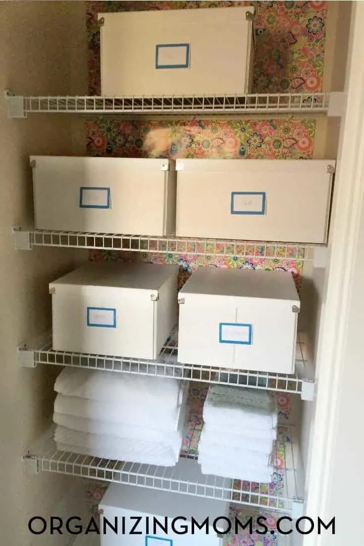 A linen closet with floral wallpaper on the back wall, storage boxes on shelves with labels and some white towels stacked neatly on the bottom shelf.