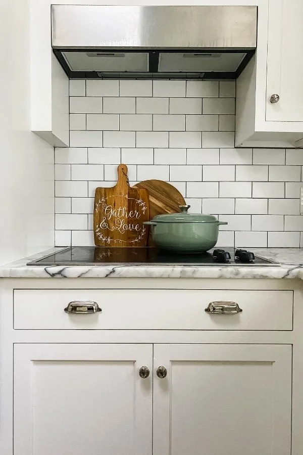 An aqua colored dutch oven on a stove top with white cabinets, white subway tile back splash and two cutting boards in the background.