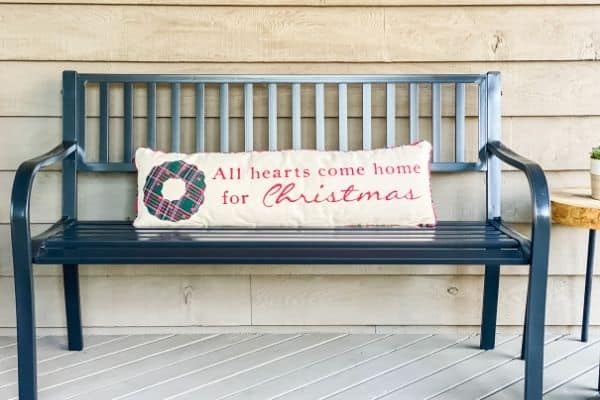 The front porch bench with a long lumbar pillow that says All hearts come home for Christmas.
