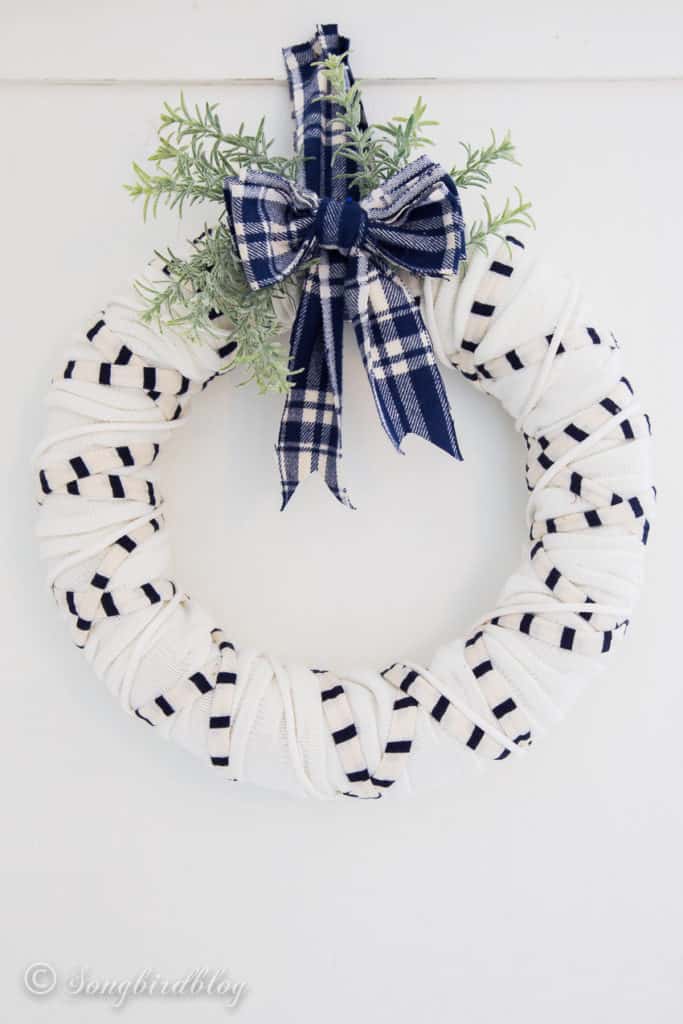 A white yarn wreath with a plaid blue and white ribbon and sprigs of evergreen.