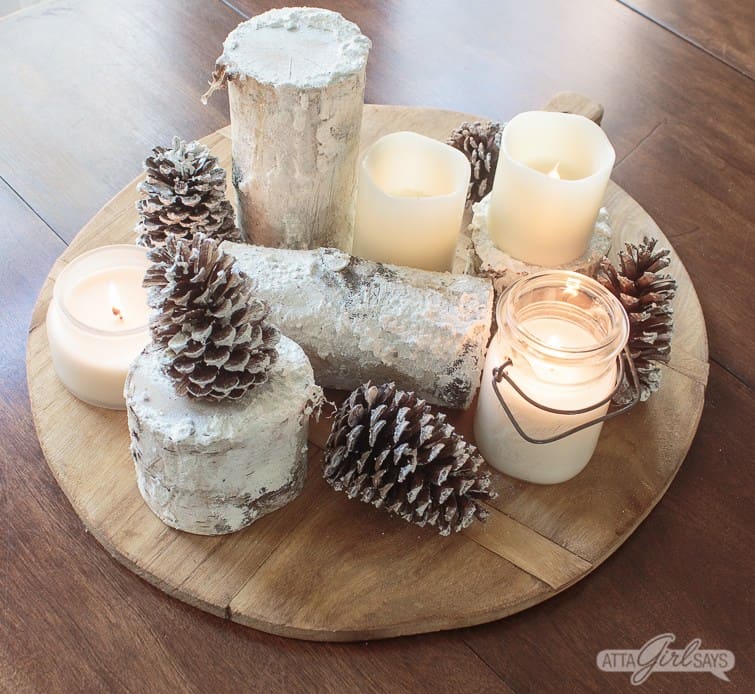 Candles, pinecones and birch logs on a wood round cutting board.