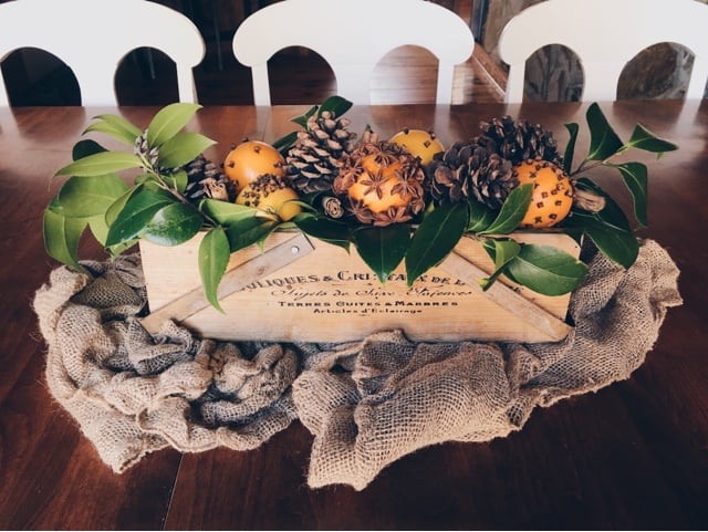 A wooden box on burlap with leaves, pine cones and oranges with spices stuck in them on a dining table.