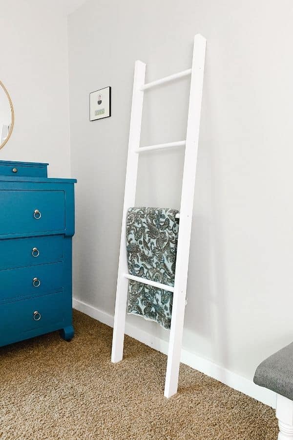 A room with a blue dresser, light gray walls and a blanket ladder painted white and a paisley blanket hanging on it.