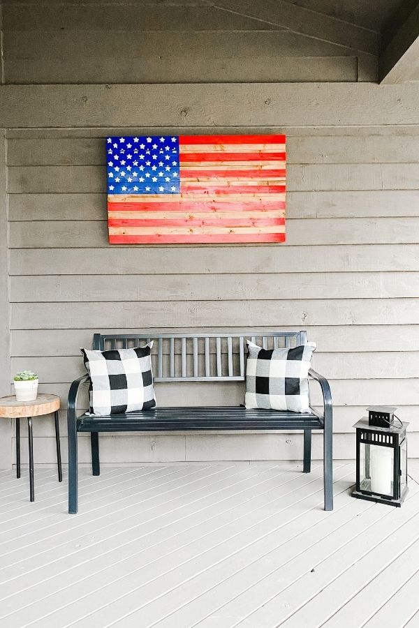 A wooden flag hanging on the wall of a small porch, a black bench with buffalo check pillows and a lantern on the floor next to it.