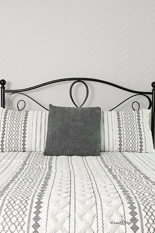 The finished herringbone stenciled wall with the black metal headboard and gray and white bedding with geometric pattern on it.