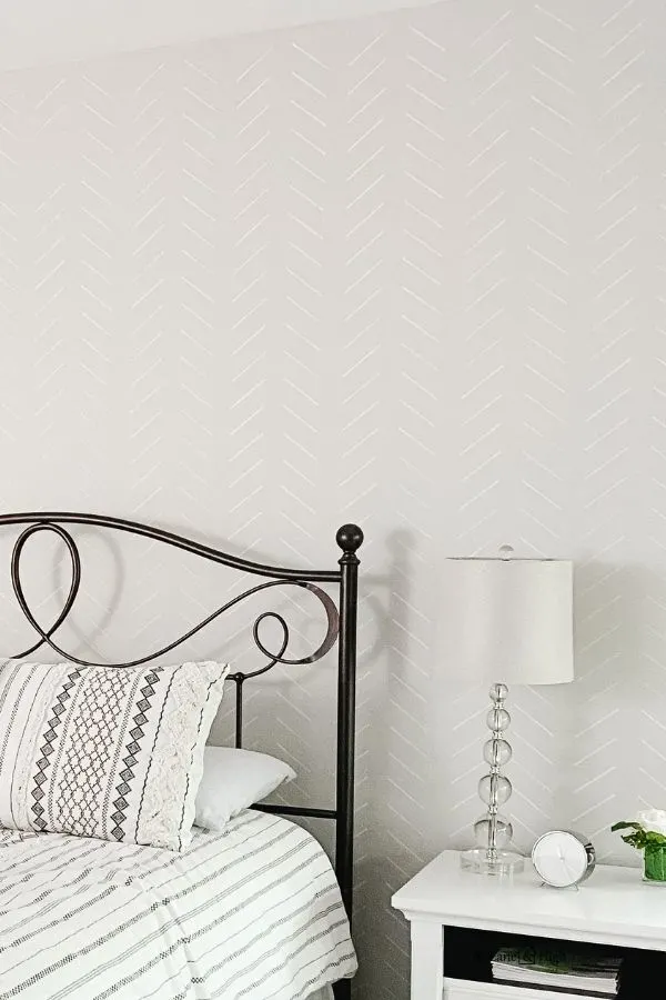 A light greige wall with white herringbone stencil in a bedroom with a black iron headboard and a white nightstand with a bubble lamp.