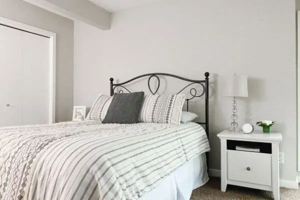 A bedroom with light gray walls, a black iron headboard and a white nightstand.