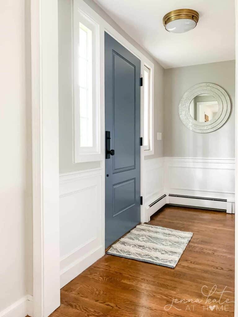 An entryway with white wainscoting, agreeable gray painted above and a blue gray painted door with black hardware.