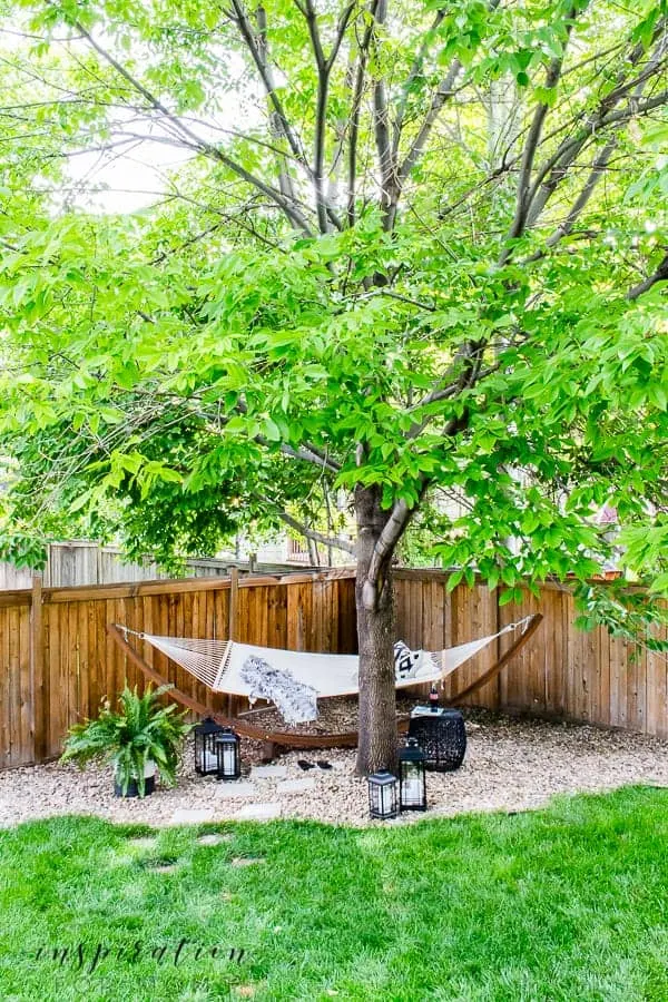 A hammock under a tree in the corner of the backyard against a fence.