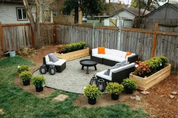 A fire pit surrounded by couches and raised garden beds with string lights on the fence behind.