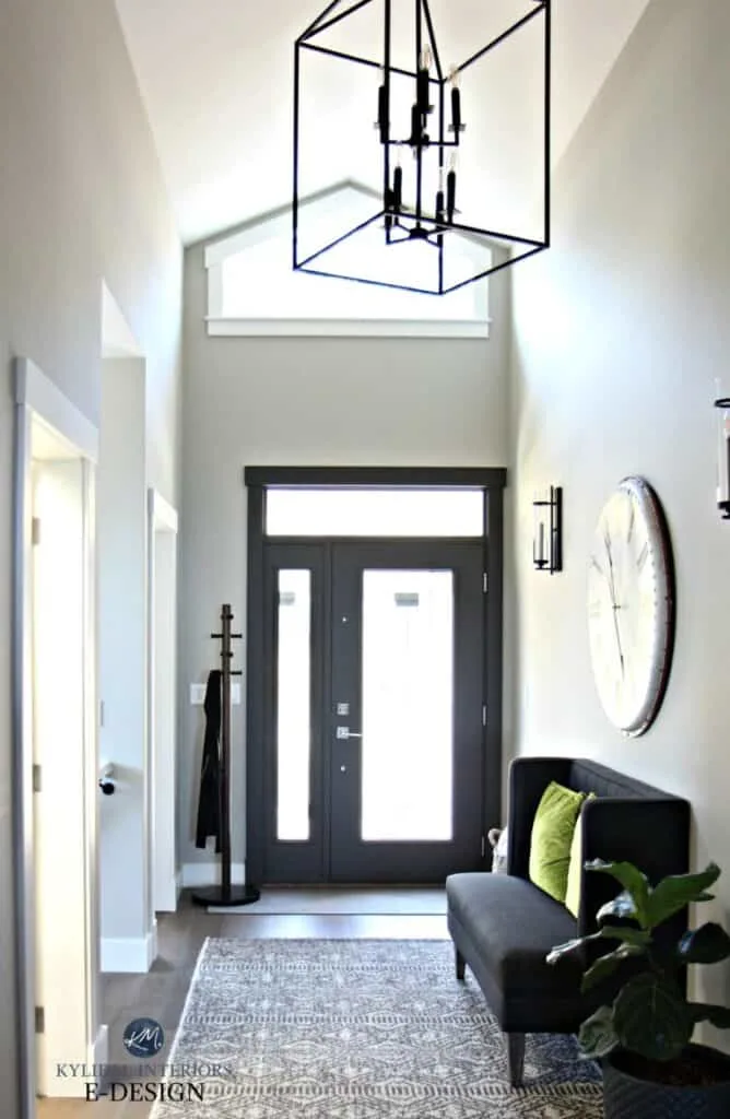 This entryway has light colored walls and a dark painted front door.  There is also a dark gray bench with a clock above.