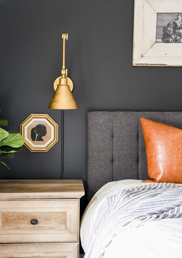 This moody master bedroom has dark painted walls with a dark gray upholstered headboard and gold wall sconce.
