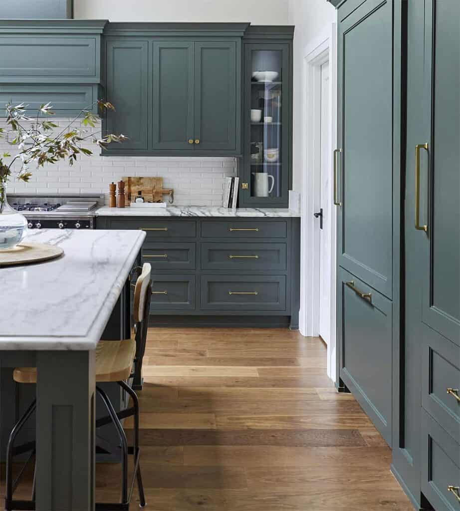 A kitchen with dark green on the cabinets, gold hardware, marble counters and hardwood floors.