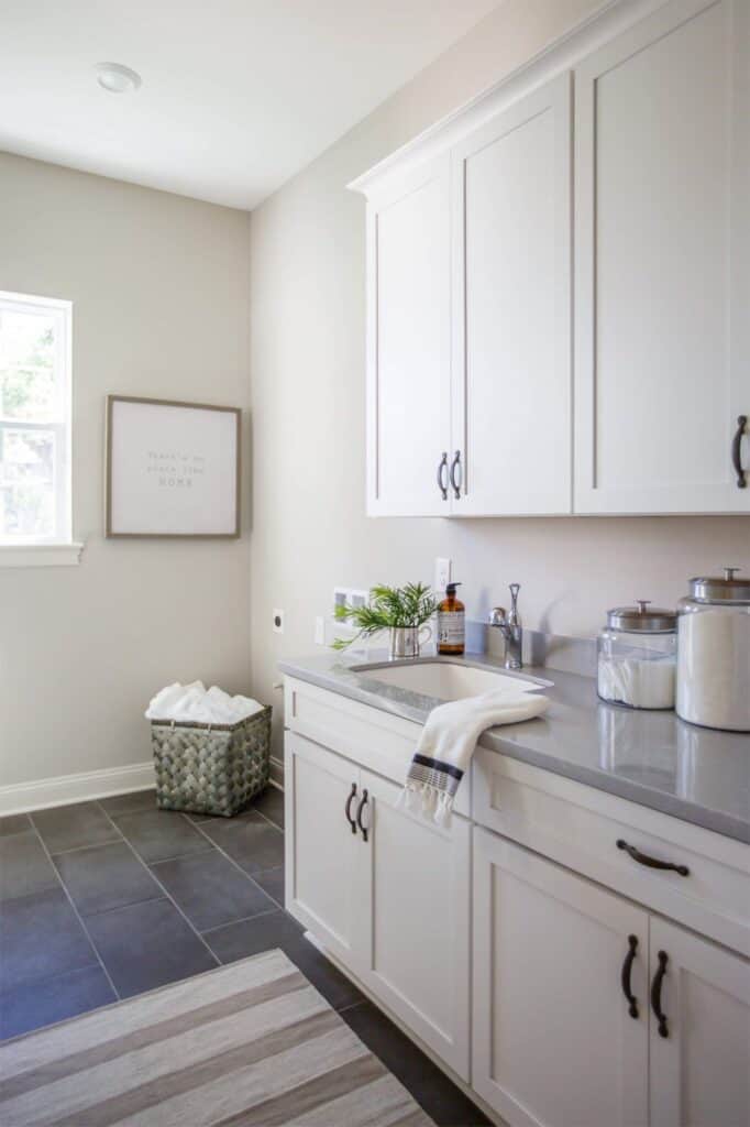 This big laundry room has dark gray tile floors, white cabinets, gray countertops and Sherwin Williams Worldly Gray on the walls.
