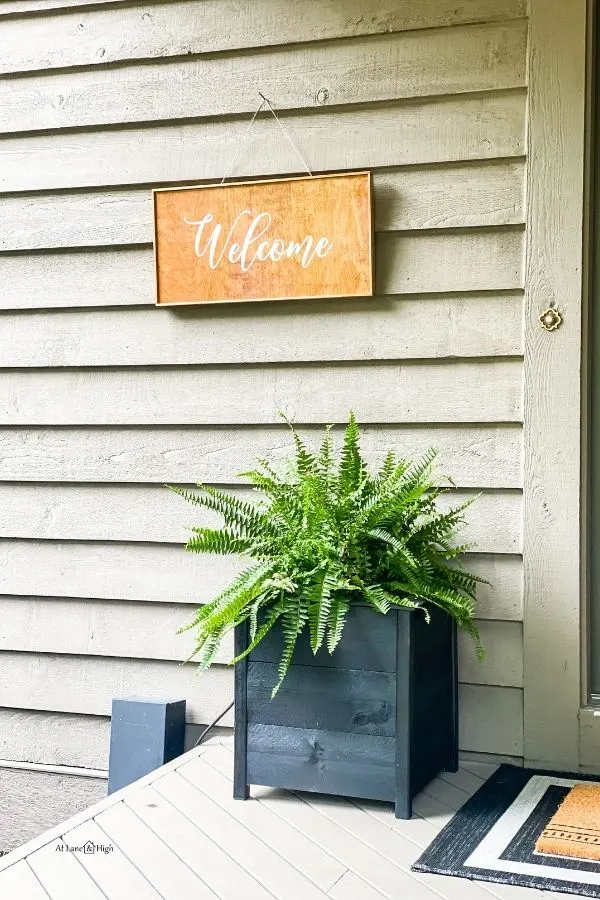 The black porch planter with a fern and a welcome sign.