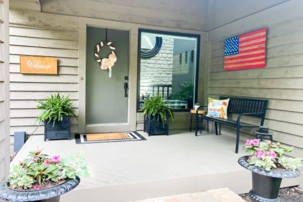 A front porch with a wooden flag, a welcome sign, planters flanking the doors with ferns in them and a back iron bench.