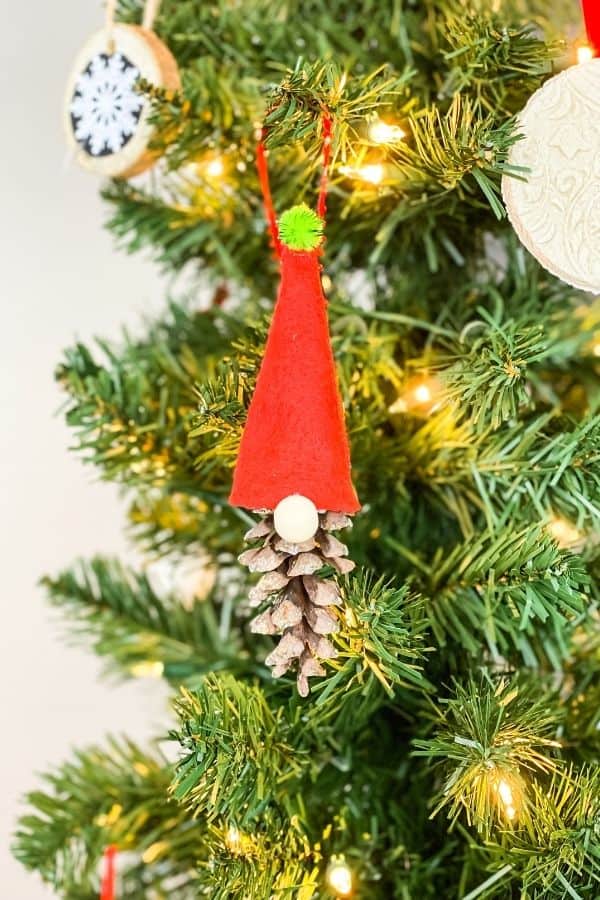 Here is a long pinecone gnome with a red had and green pompom hanging on a Christmas tree.