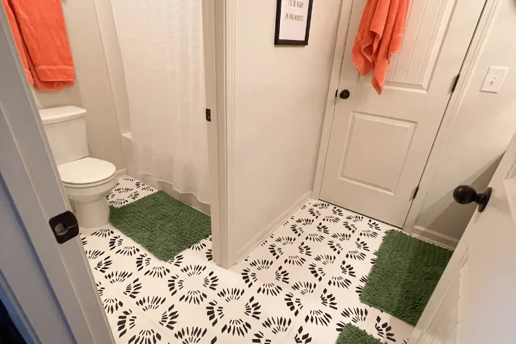 A bathroom with painted floor tile and a stencil in black and white with green bath rugs and orange towels.