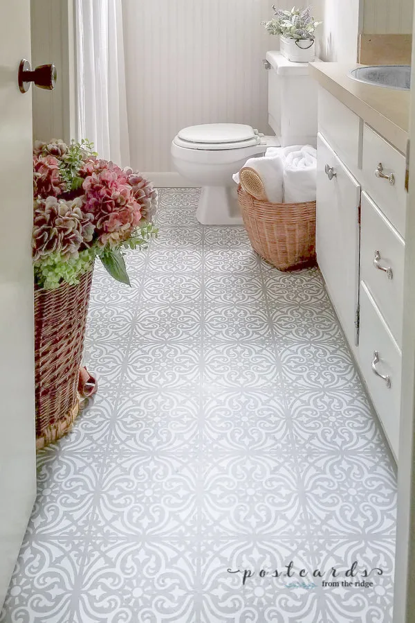 A bathroom with linoleum floors that have been painted using a stencil in gray and white and a basket with flowers sitting on the wall opposite the vanity.