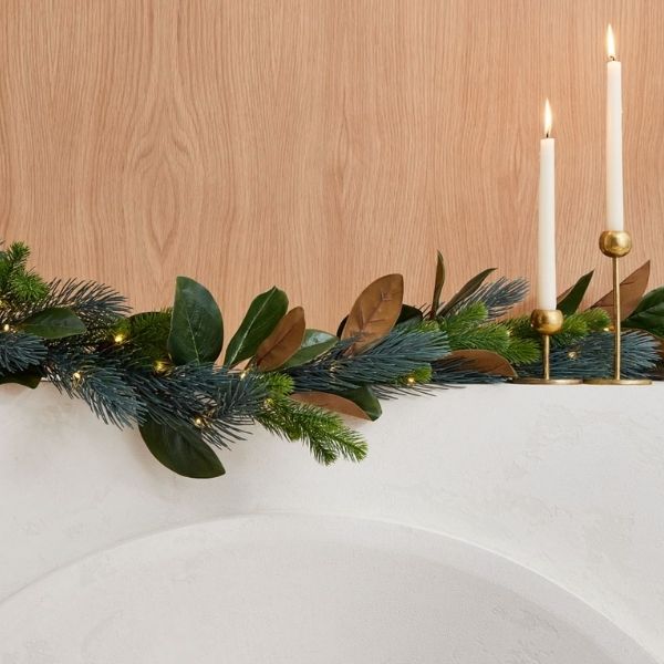A magnolia wreath on a white mantel with little white lights and two white candles on gold candle sticks.