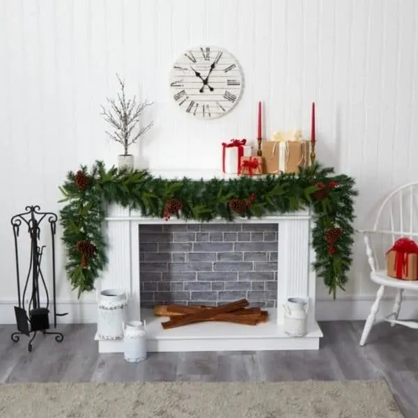 A very full garland with pinecones hung on a white mantel with wrapped presents, red candles and a clock hung on the wall above the mantel.