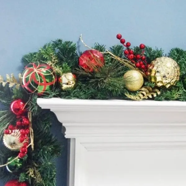 A garland on top of a white mantel with red berries and ball ornaments in red, green and gold.