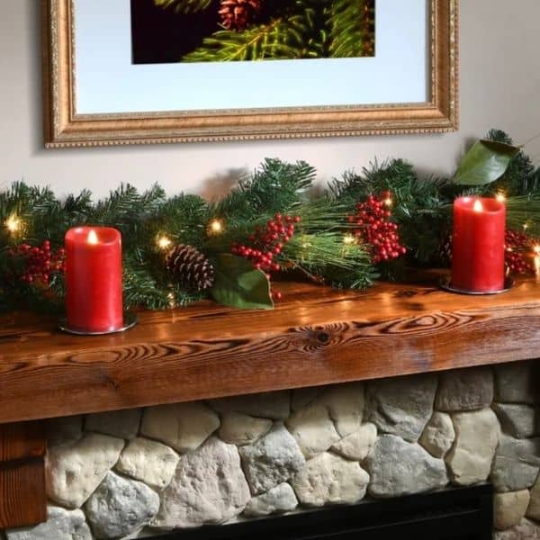 A garland with red berries and pinecones on a wood mantel of a stone fireplace with red candles.