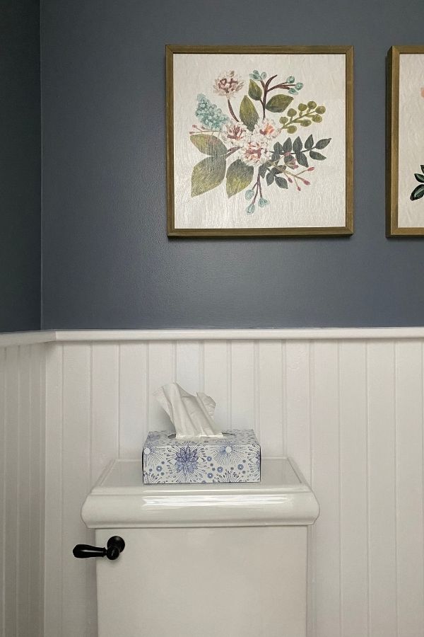 tongue and groove wainscoting in a powder room with dark gray blue walls above and floral artwork.