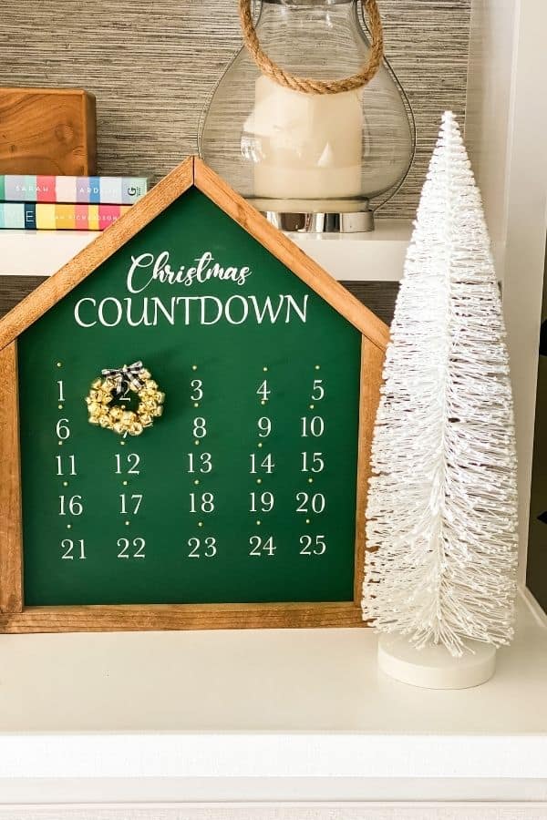 A green advent calendar with white writing, a wood frame and a gold jingle bell wreath for each day.